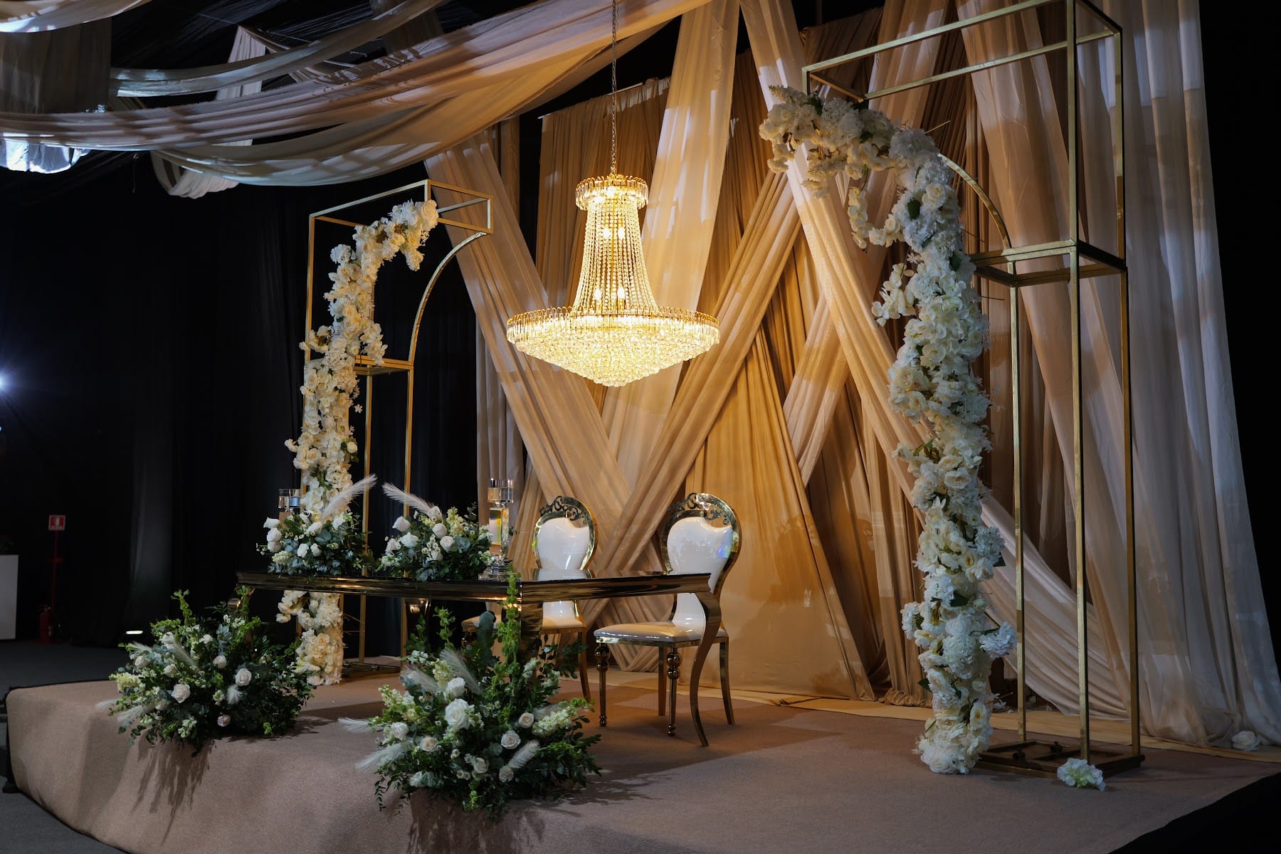 Escenario de boda con telas doradas, candelabro y arreglos de flores blancas