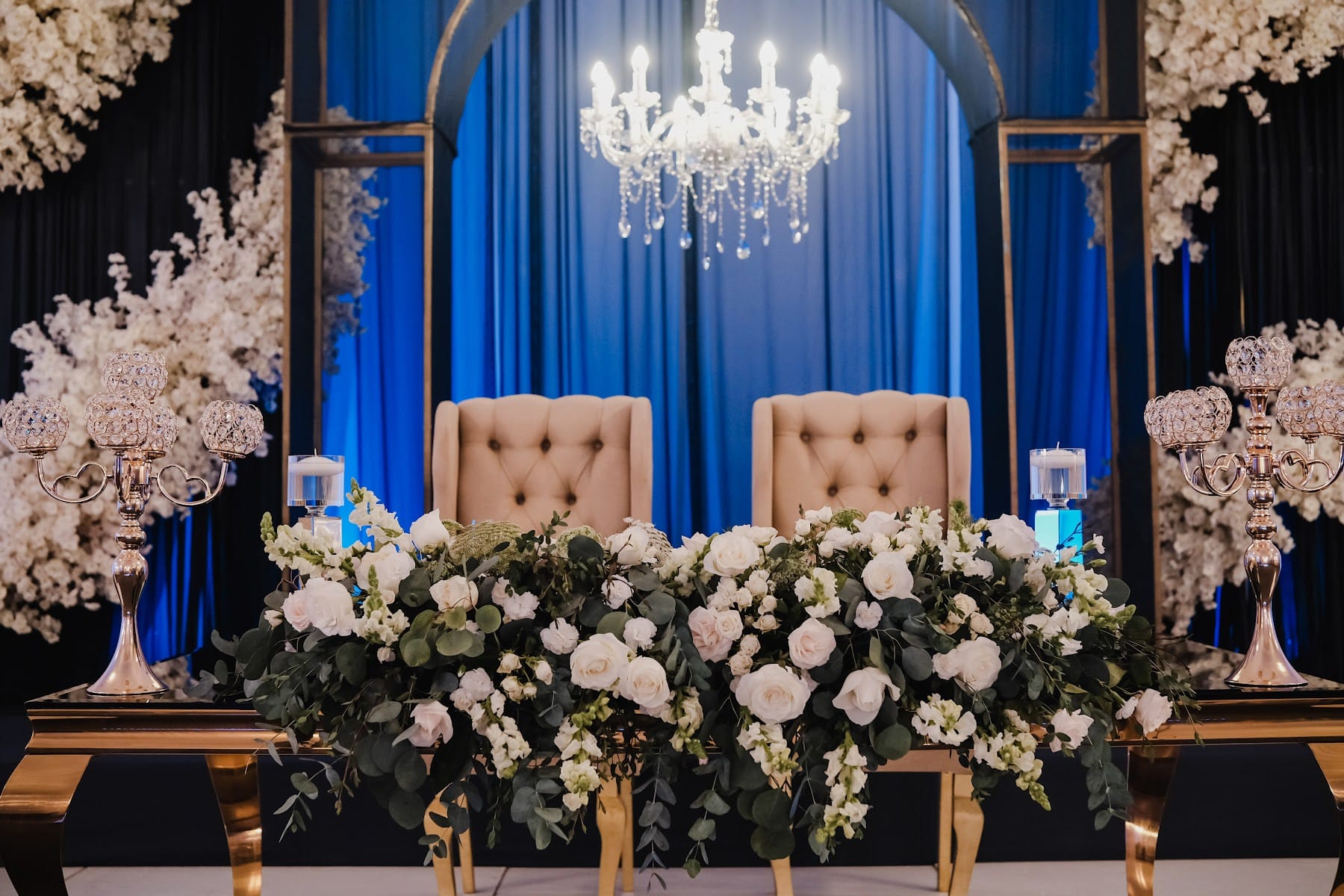 Mesa de novios con sillas capitonadas y arreglo de rosas blancas sobre fondo azul