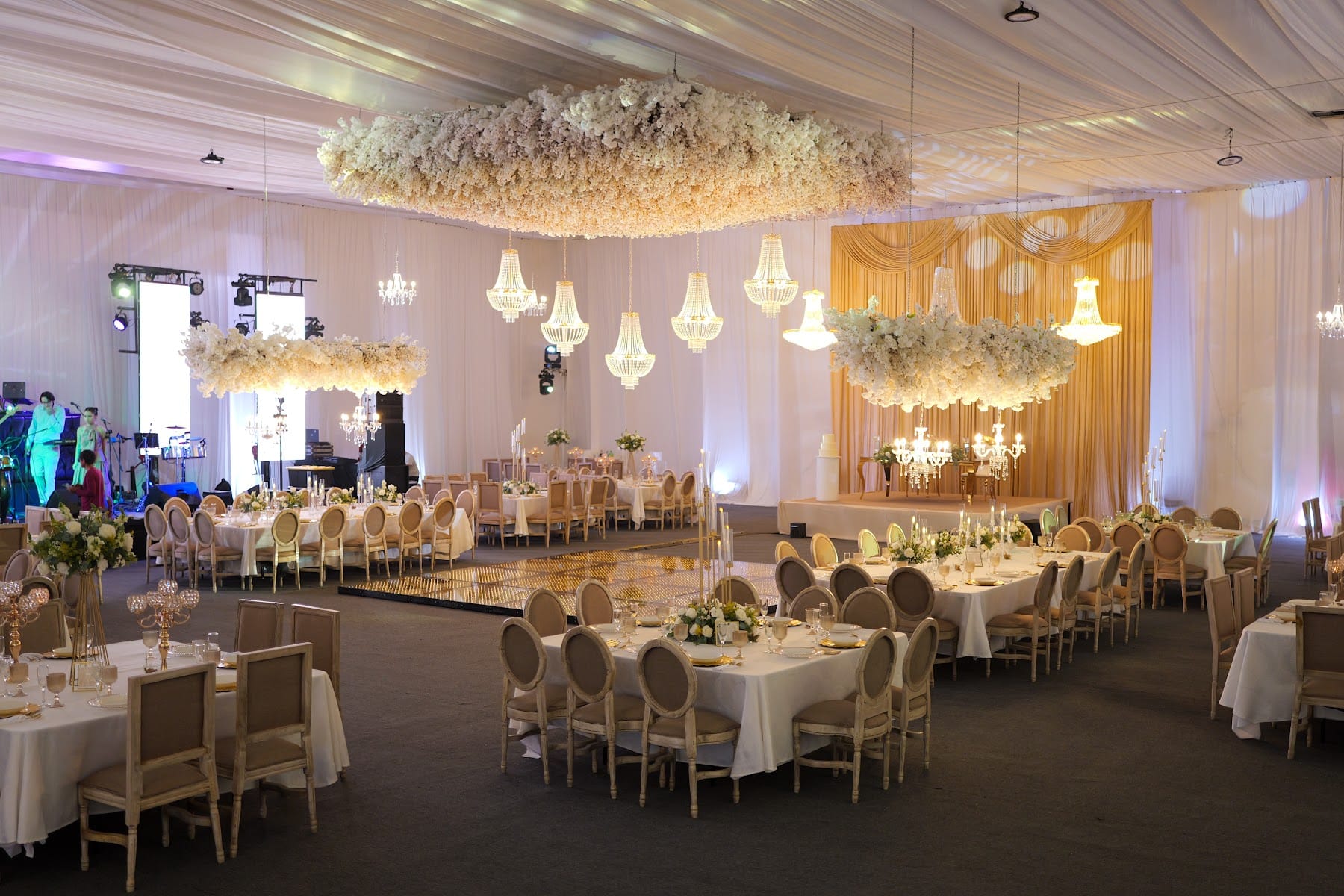 Salón de boda con entelado blanco, nubes de flores y candiles de cristal sobre mesas largas