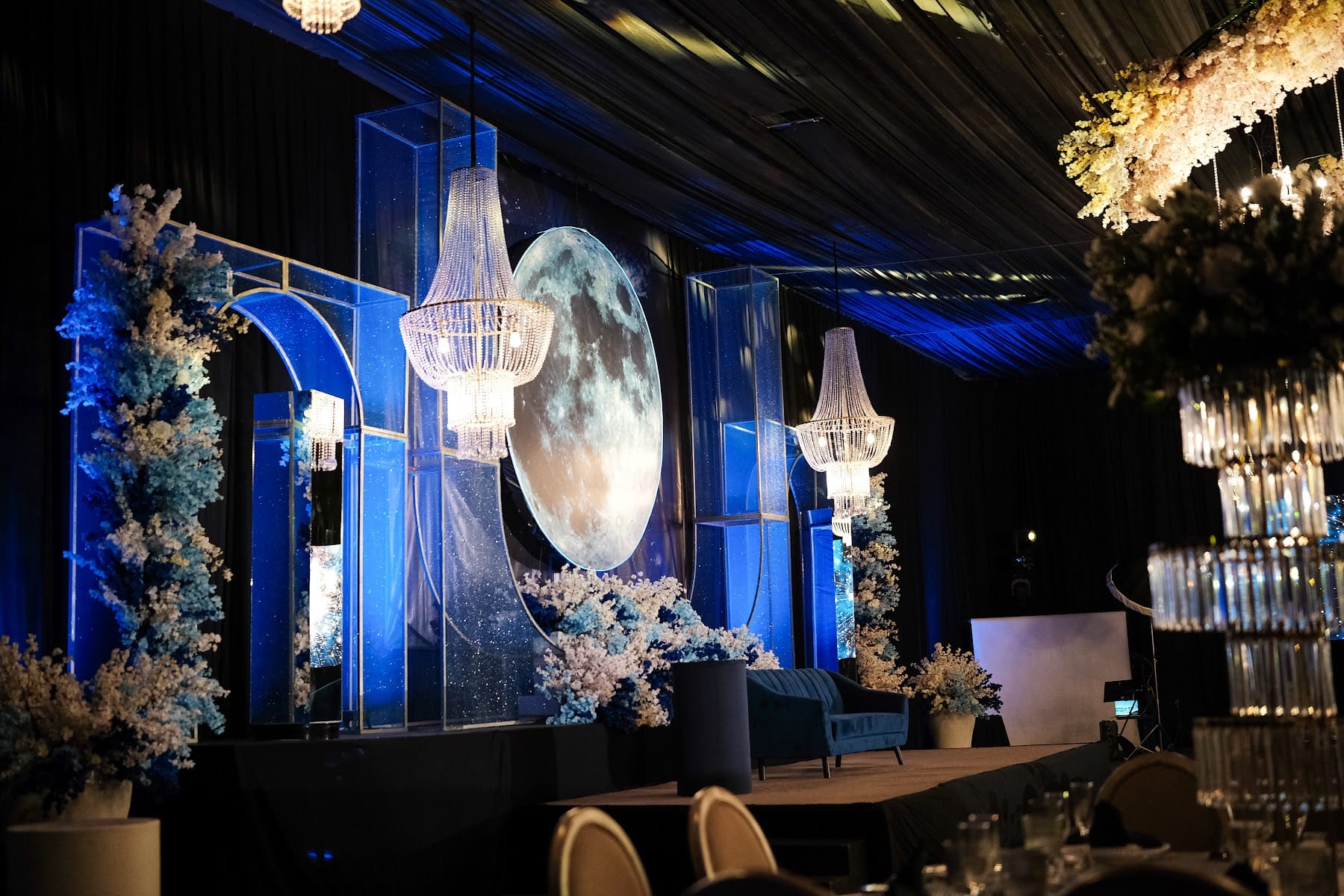 Escenario con luna gigante, arcos de cristal y flores blancas en luz azul para los XV años de Camila
