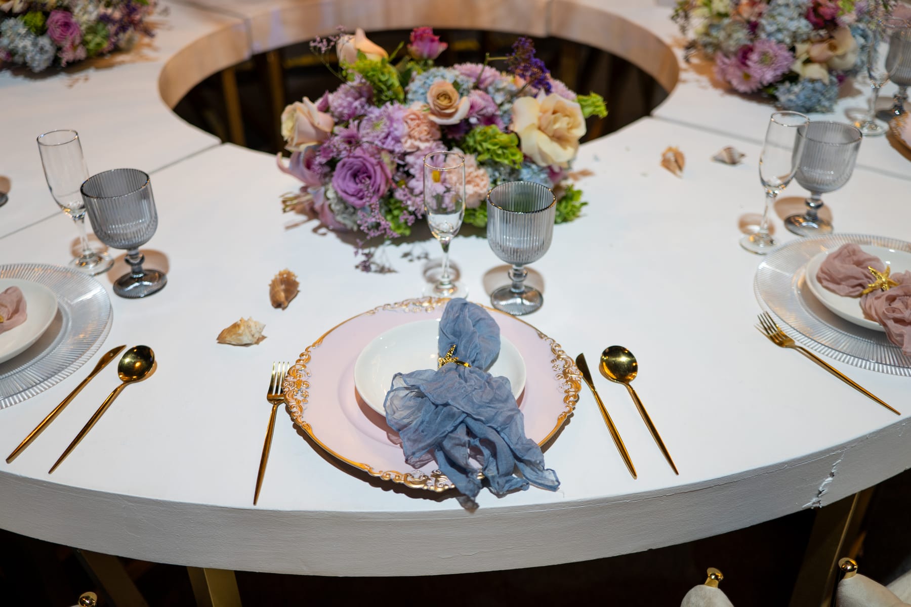 Mesa curva blanca con flores, conchas, servilletas azules y cubiertos dorados