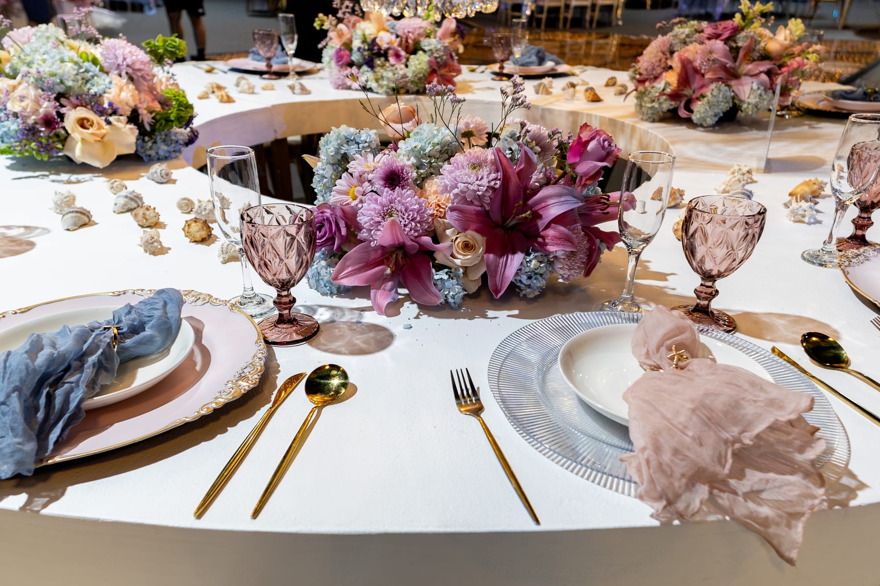 Mesa blanca con centro de flores lila y rosa, copas de color y cubiertos dorados